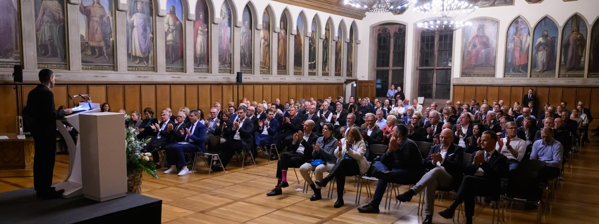 Ein Man steht an einem Rednerpult und spricht zu mehr als hundert Personen, die in einem Festsaal auf Stuhlreihen sitzen