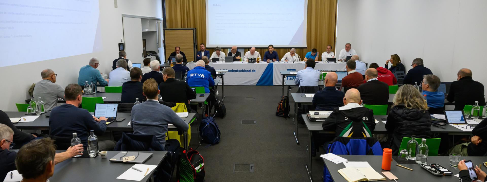 Viele Menschen sitzen in Stuhlreihen, weiter hinten sitzen Personen, die die Sitzung halten