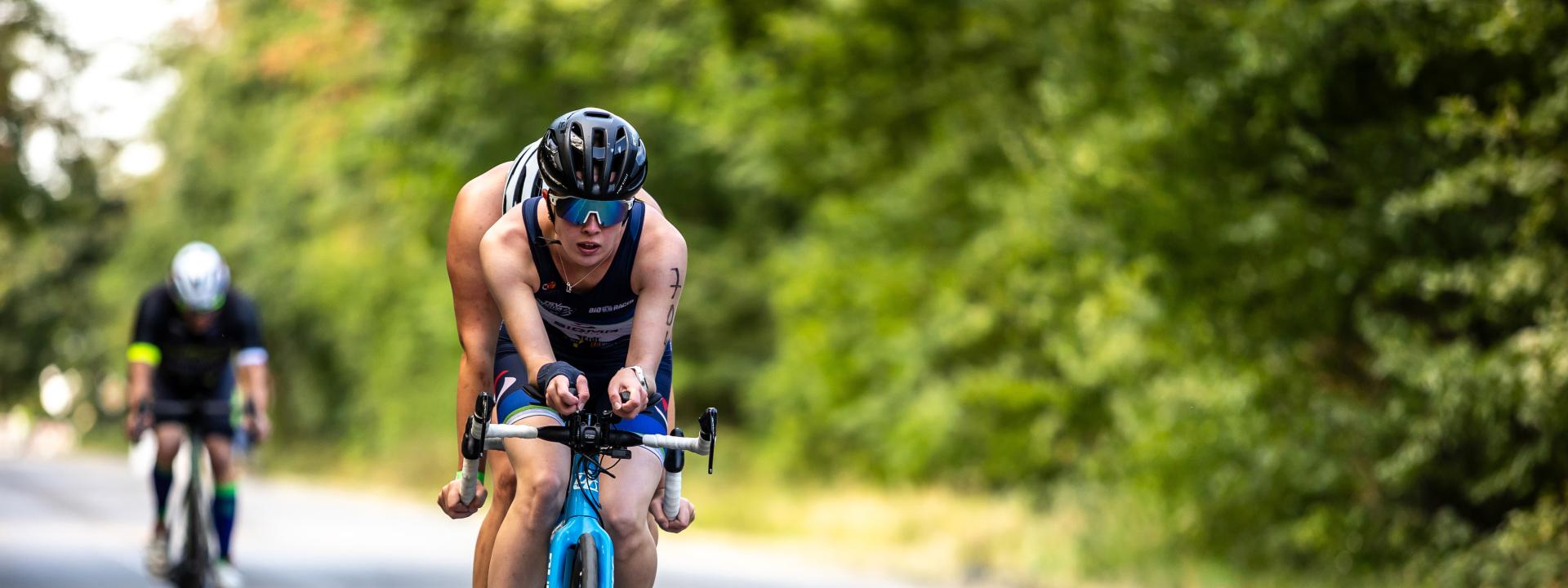 Ein Tandem fährt auf einer Straße, dahinter fährt ein einzelner Radfahrer