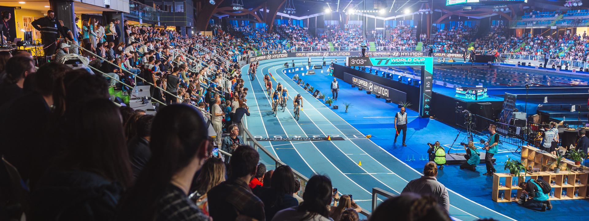 Mehrere Triathleten fahren im Rahmen eines Indoor-Events auf dem Rundkurs einer Halle an einer großen Zuschauermenge vorbei