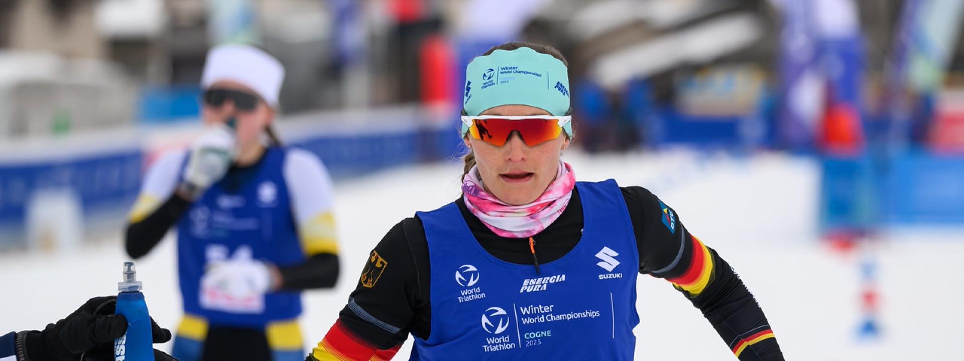 Eine Wintertriathletin in deutschem Rennanzug bekommt auf der Laufstrecke eine Trinkflasche gereicht
