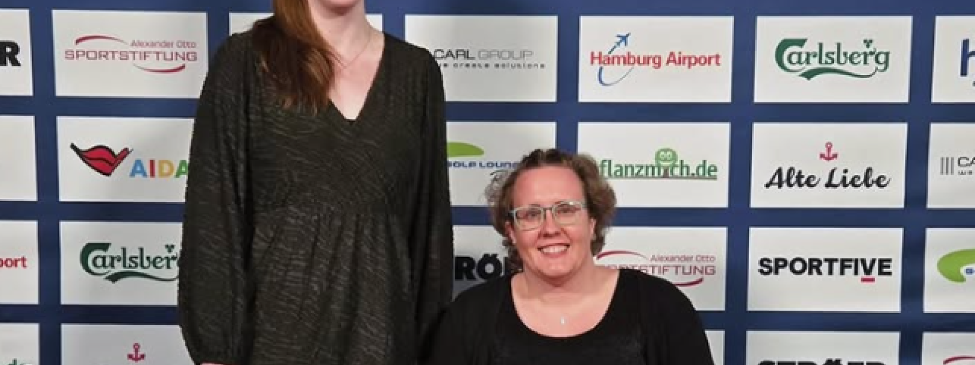 Eine Frau im Rollstuhl und eine Frau neben ihr stehend werden vor einer Sponsorenwand im Hintergrund portraitiert