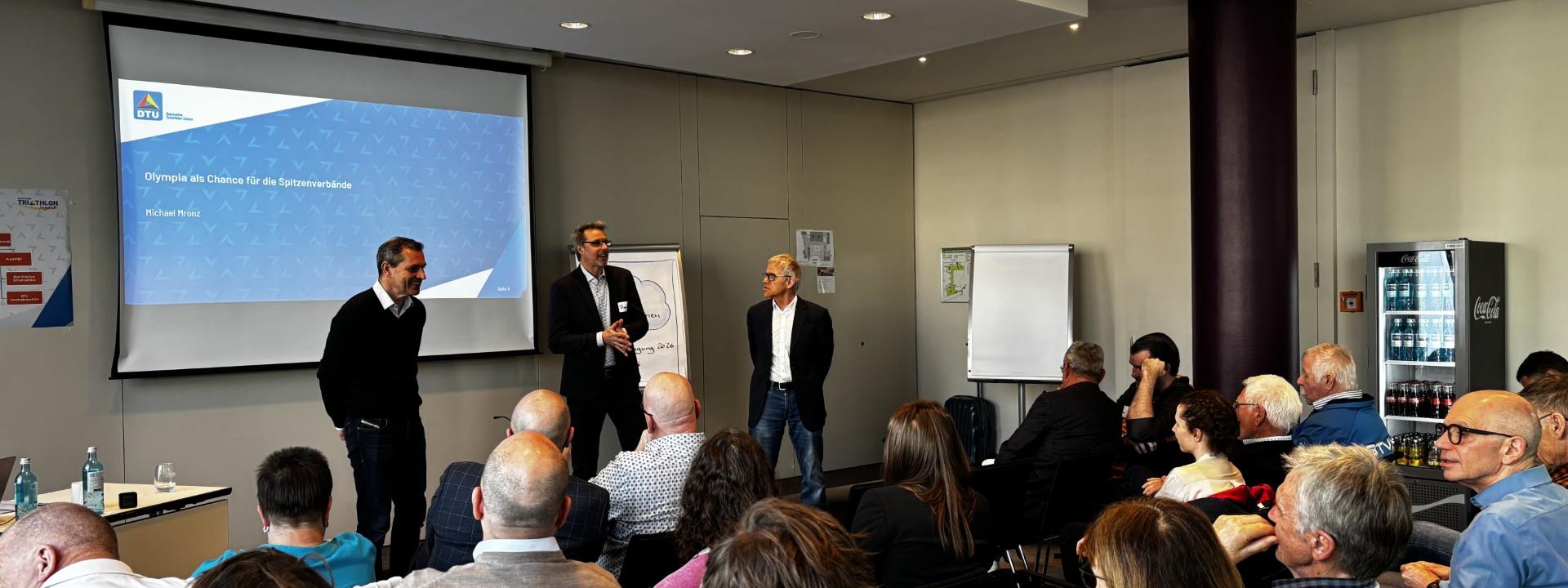 Drei Männer sprechen im Rahmen einer Präsentation zu vielen Menschen, die auf Stuhlreihen sitzen und zuhören