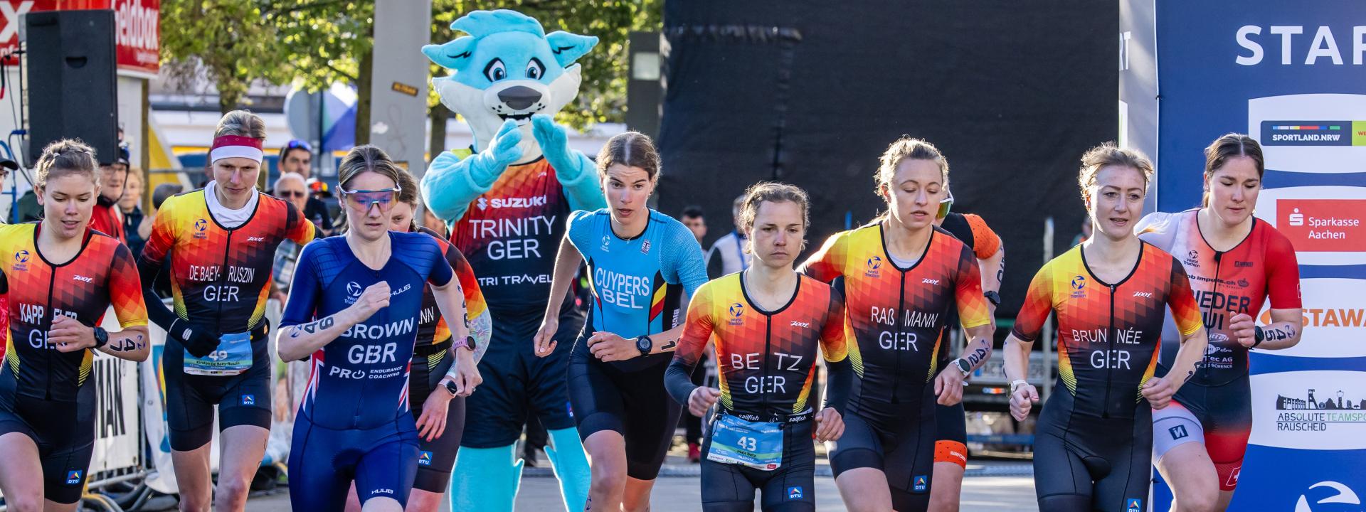 Athletinnen der Duathlon WM in Alsdorf laufen an der Startlinie los, im Hintergrund Maskottchen Trinity