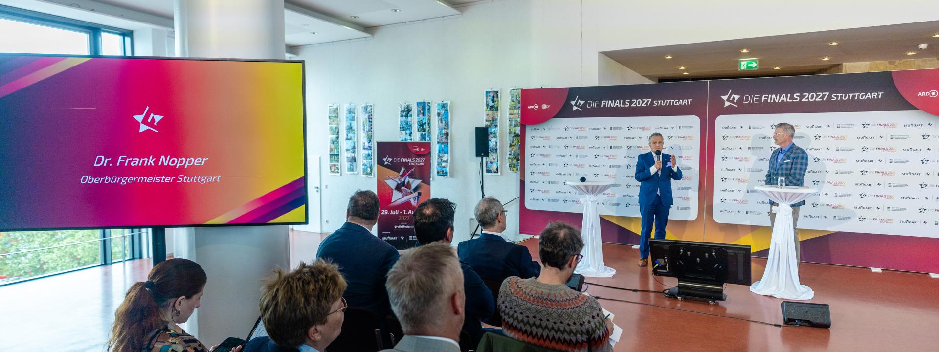 Zwei Männer unterhalten sich vor einer Presserückwand im Rahmen einer Pressekonferenz. Im Vordergrund sieht man mehrere Menschen, die auf Stühlen sitzend interessiert zuhören
