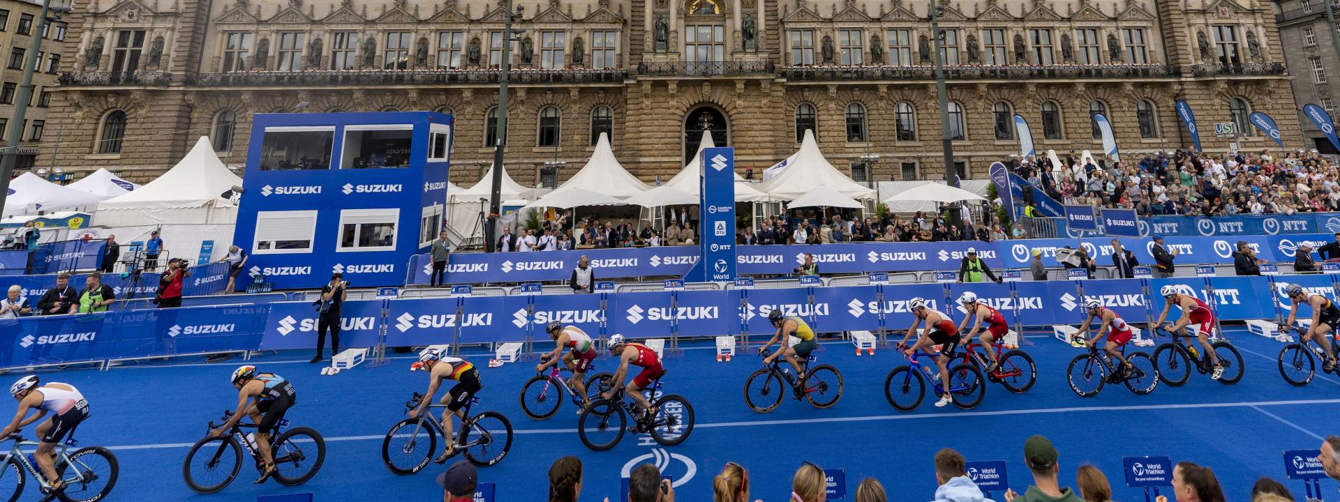 Zahlreiche Triathleten fahren auf dem Rad wie an einer Perlenschnur aufgereiht hintereinander durch die Wechselzone. Im Hintergrund ist das Hamburger Rathaus prominent zu sehen