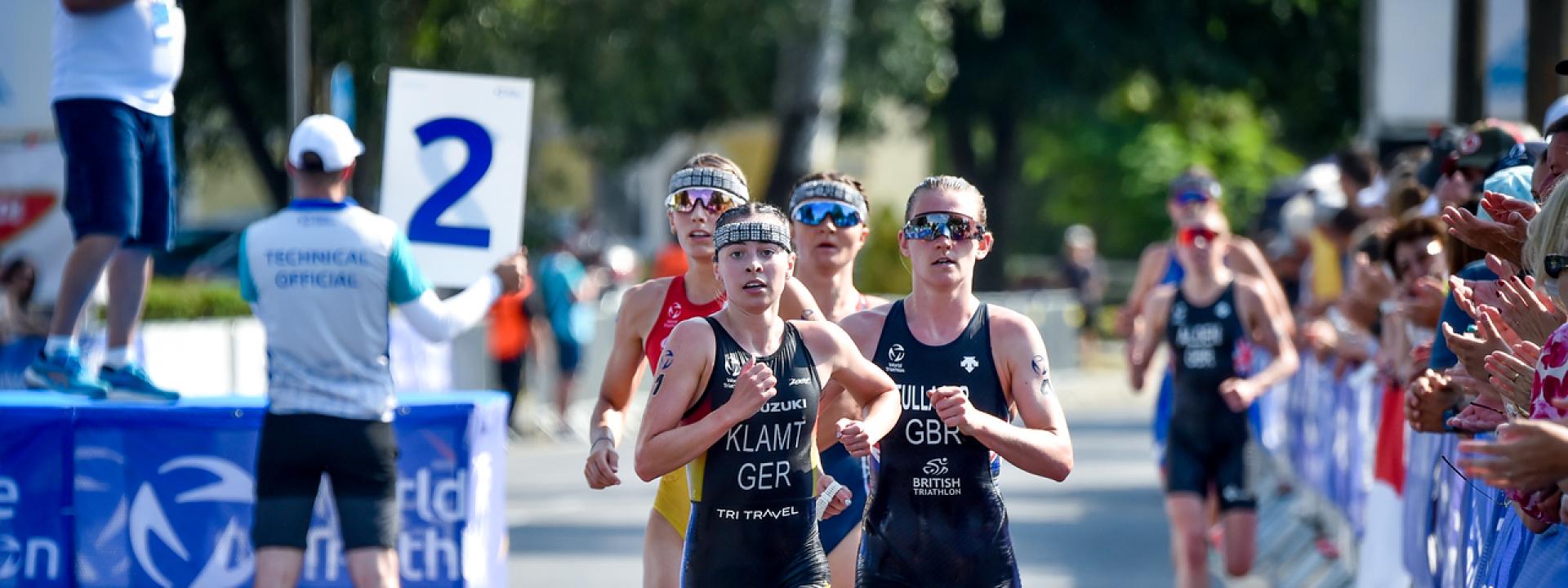 Mehrere Triathletinnen laufen in einer Gruppe über die Asphaltstrecke einer Triathlonveranstaltung. Im Hintergrund jubeln Menschen.