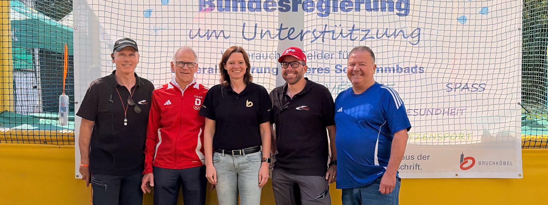 Vier Männer und eine Frau posieren vor einem Plakat, das um Unterstützung zur Sanierung des Bruchköbler Schwimmbads bittet