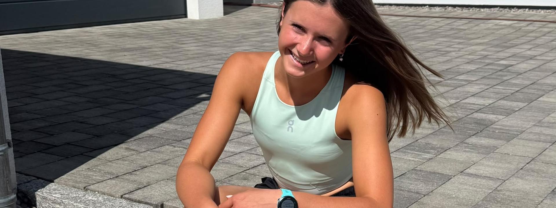 Junge Triathletin sitzt auf Pflasterstein und lacht in die Kamera