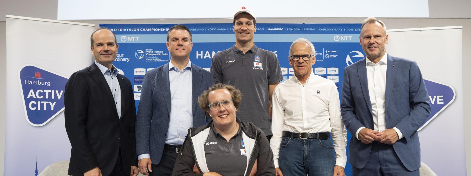 Fünf Männer posieren gemeinsam mit einer Rollstuhlfahererin für ein Foto auf der Bühne der Pressekonferenz