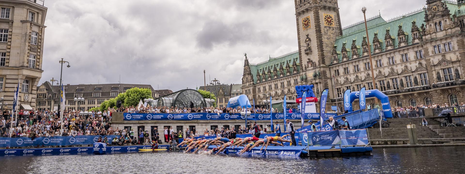Rund 15 Triathleten springen bei einem Start von einem künstlichen Steg aus ins Wasser