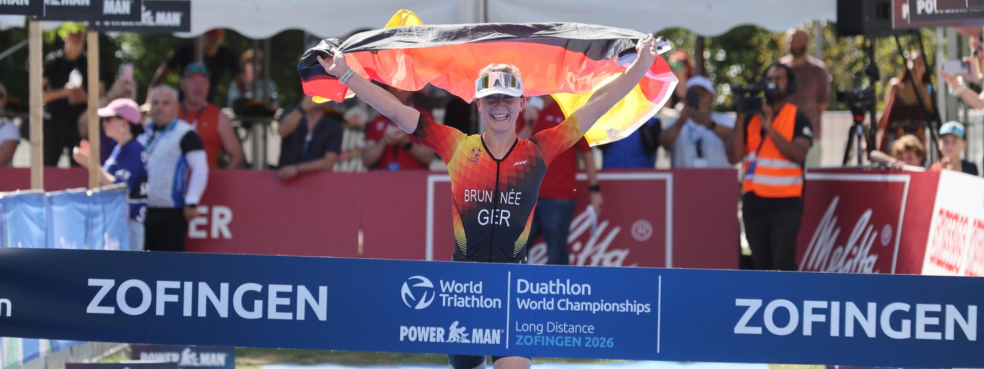 Eine Sportlerin in deutschem Dress läuft freudestrahlend mit der Deutschlandfahne in der Hand dem Sieg bei einem WM-Rennen entgegen