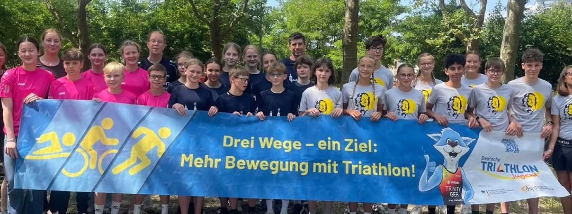 Mehrere Menschen stehen hinter einem Banner mit dem Slogan: "Drei Wege - ein Ziel: Mehr Bewegung mit Triathlon!"