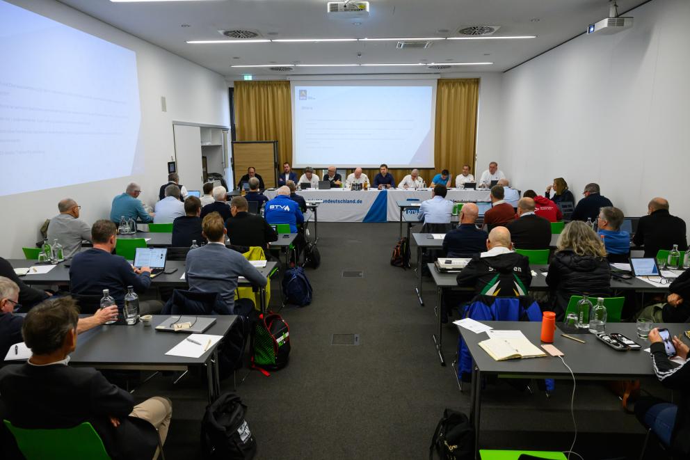 Viele Menschen sitzen in Stuhlreihen, weiter hinten sitzen Personen, die die Sitzung halten