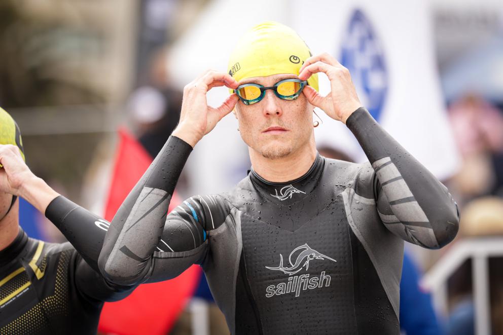 Ein Mann im Neoprenanzug und mit Badekappe auf dem Kopf bereitet sich auf einen Triathlon vor, indem er die Schwimmbrille aufzieht