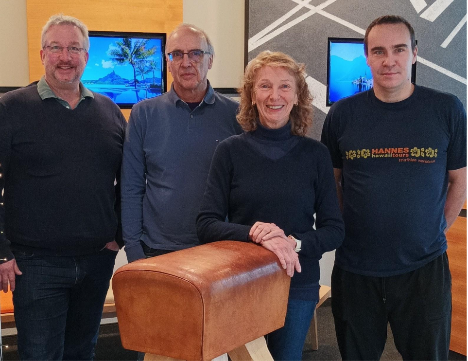 Drei Männer und eine Frau posieren an einem kleinen Turngerät, dem Turnpferd, für ein Foto