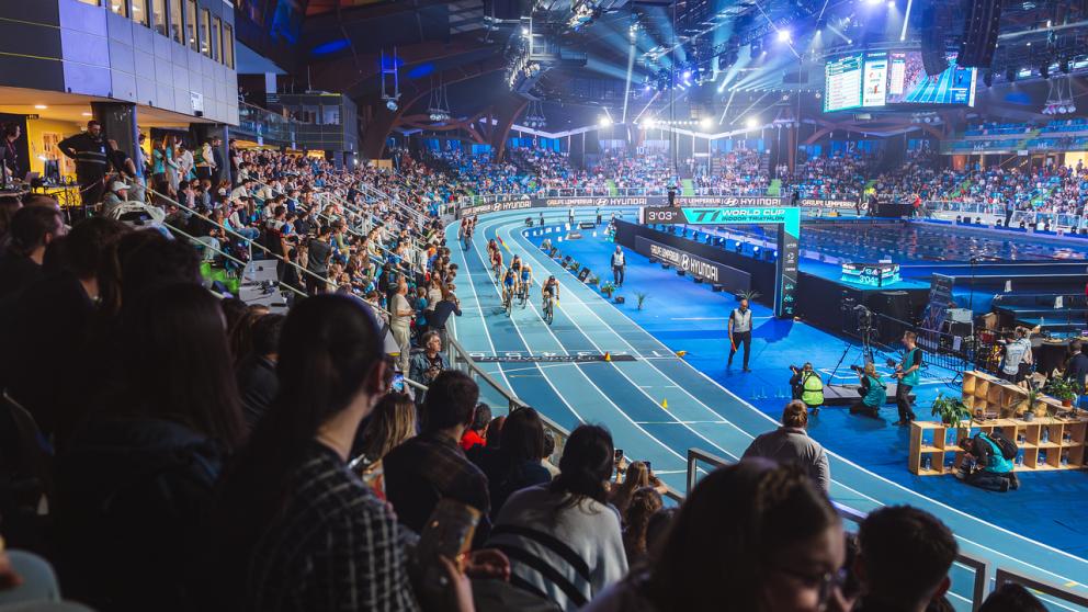 Mehrere Triathleten fahren im Rahmen eines Indoor-Events auf dem Rundkurs einer Halle an einer großen Zuschauermenge vorbei