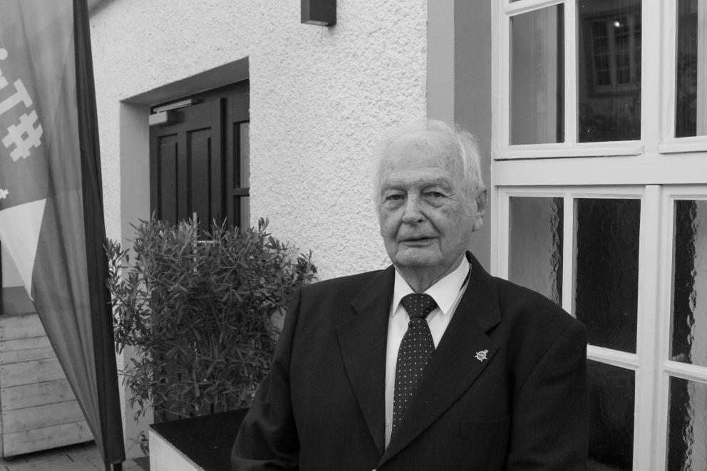 Ein älterer Mann steht vor einer Fensterfront und schaut in die Kamera