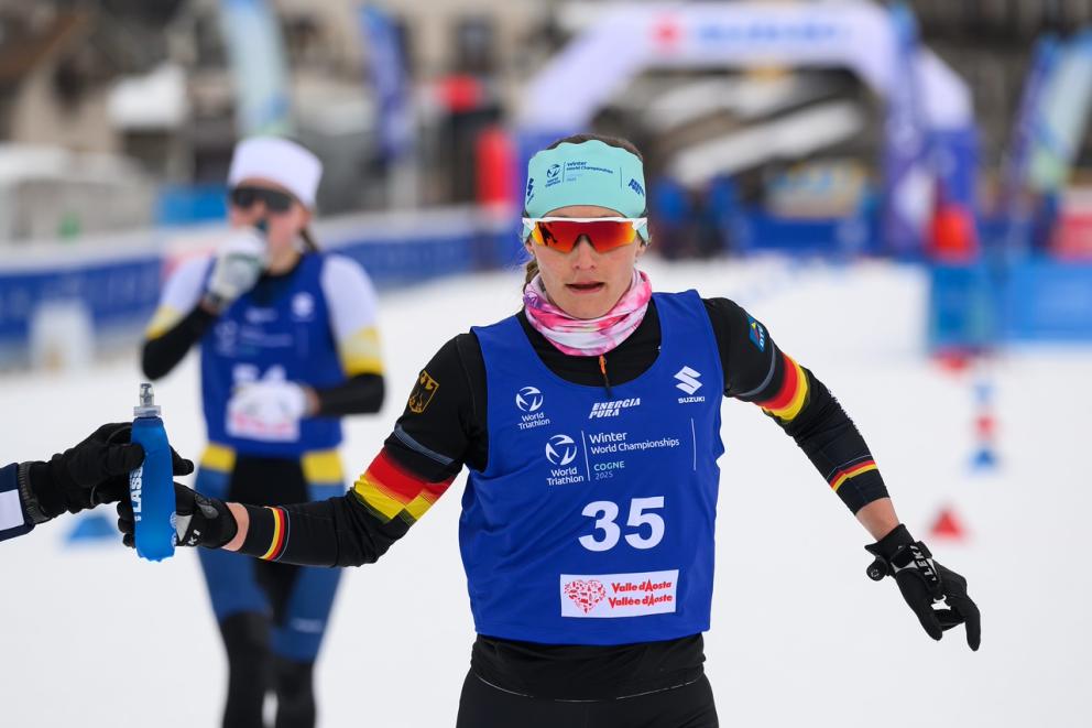 Eine Wintertriathletin in deutschem Rennanzug bekommt auf der Laufstrecke eine Trinkflasche gereicht