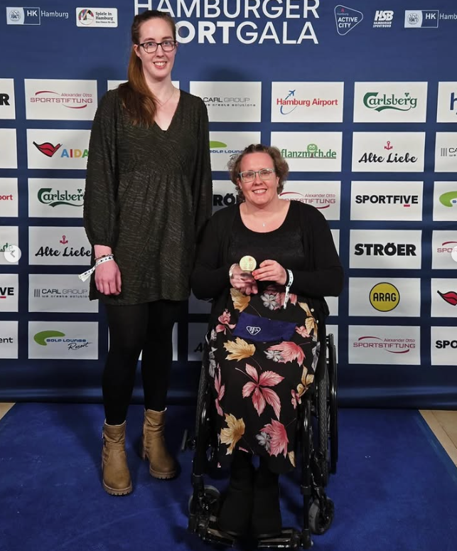 Eine Frau im Rollstuhl und eine Frau neben ihr stehend werden vor einer Sponsorenwand im Hintergrund portraitiert