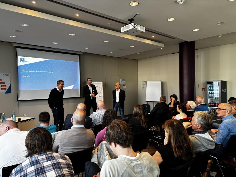 Drei Männer sprechen im Rahmen einer Präsentation zu vielen Menschen, die auf Stuhlreihen sitzen und zuhören