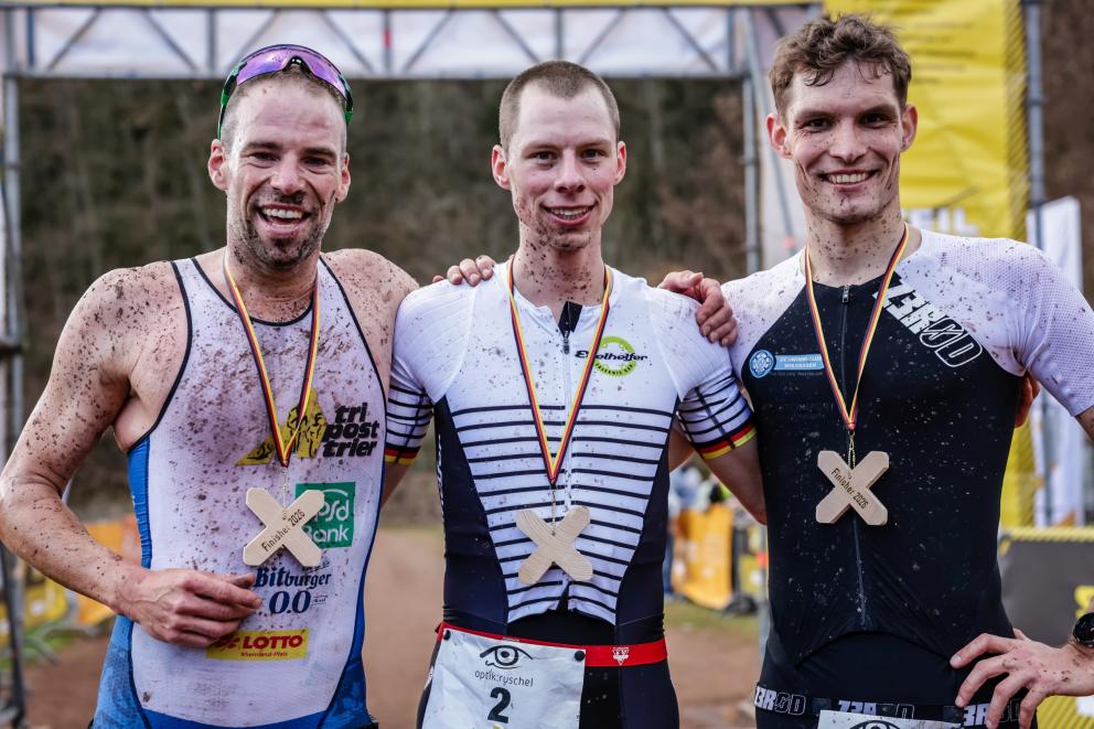 Drei Männer mit matschverspritzen Gesicht strahlen in die Kamera