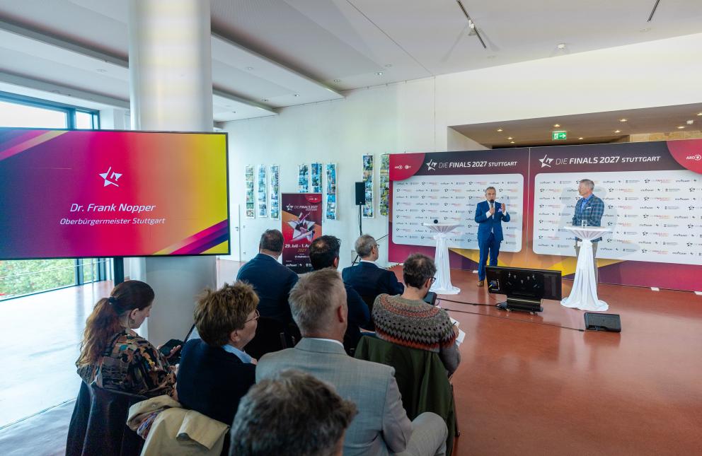 Zwei Männer unterhalten sich vor einer Presserückwand im Rahmen einer Pressekonferenz. Im Vordergrund sieht man mehrere Menschen, die auf Stühlen sitzend interessiert zuhören