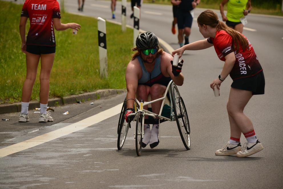 Ein Rollstuhlfahrer passiert im Rahmen eines Triathlon-Wettkampfes eine Verpflegungsstelle, an der eine junge Frau ihm Wasser zureicht