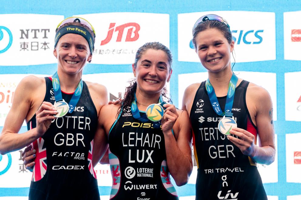 Drei Triathletinnen zeigen strahlend ihre Medaillen auf dem Podest in die Kamera