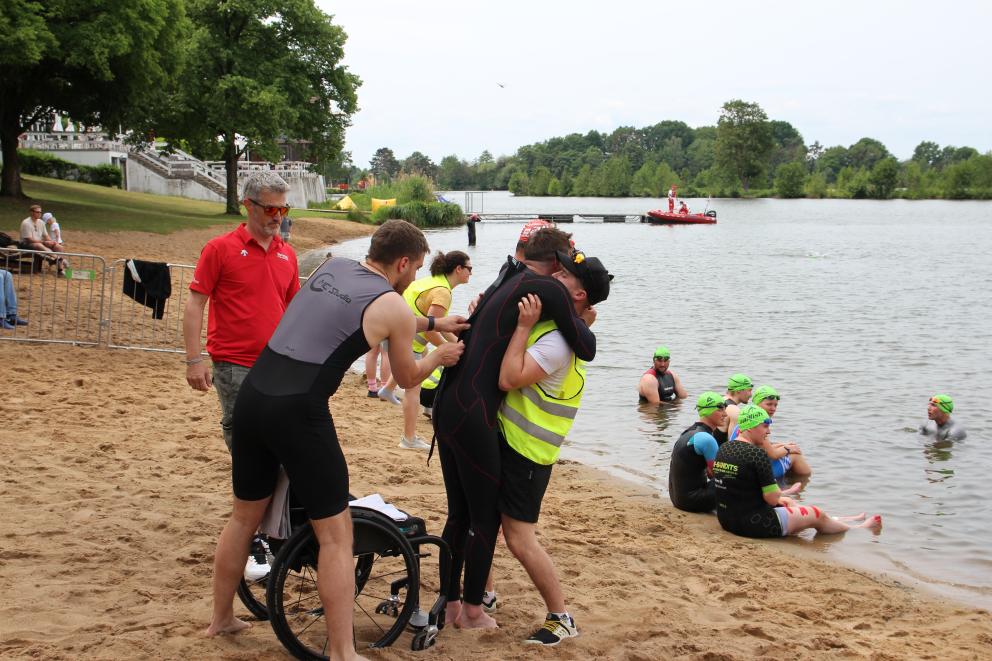 Ein Mann hebt einen Para Triathleten wenige Meter vor dem Seezugang auf einem Sandstrand aus dem Rollstuhl. Ein zweiter Mann unterstützt leicht,  ein weiterer beobachtet die Szenerie