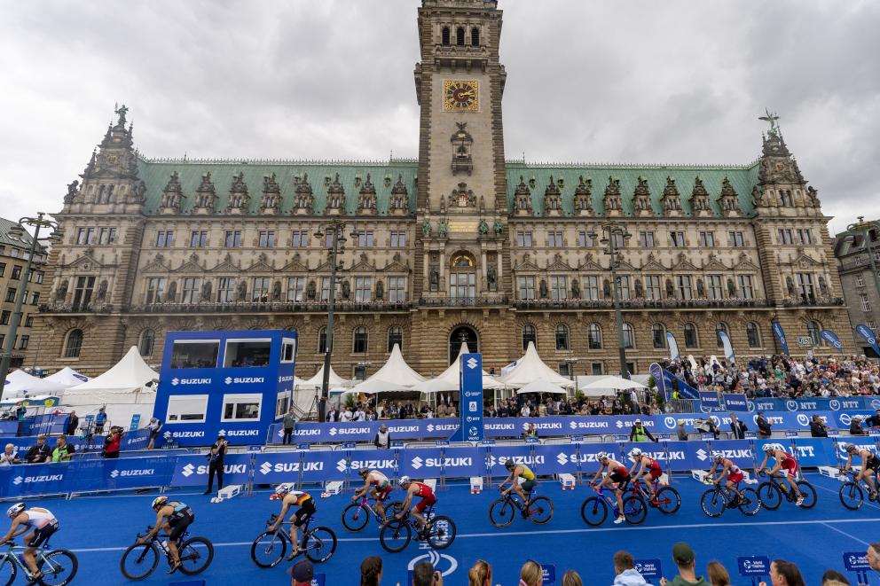 Zahlreiche Triathleten fahren auf dem Rad wie an einer Perlenschnur aufgereiht hintereinander durch die Wechselzone. Im Hintergrund ist das Hamburger Rathaus prominent zu sehen