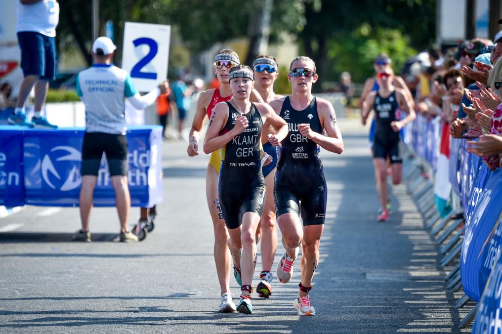 Mehrere Triathletinnen laufen in einer Gruppe über die Asphaltstrecke einer Triathlonveranstaltung. Im Hintergrund jubeln Menschen.