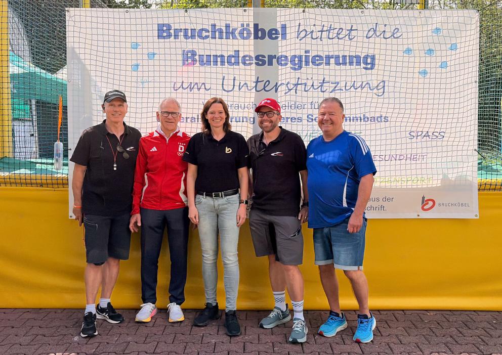 Vier Männer und eine Frau posieren vor einem Plakat, das um Unterstützung zur Sanierung des Bruchköbler Schwimmbads bittet