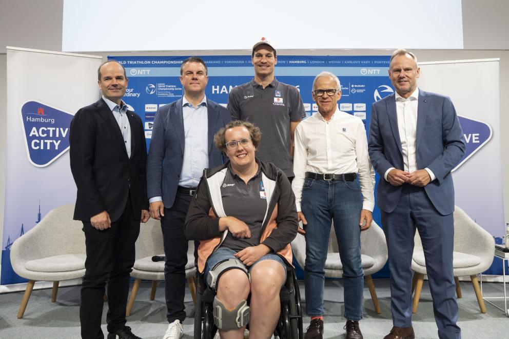 Fünf Männer posieren gemeinsam mit einer Rollstuhlfahererin für ein Foto auf der Bühne der Pressekonferenz