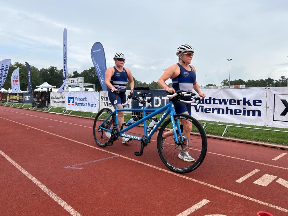 Eine sehbeinträchtige Sportlerin und ihr Guide schieben gemeinsam ein Tandem aus der Wechselzone eines Triathlons