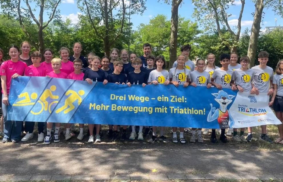 Mehrere Menschen stehen hinter einem Banner mit dem Slogan: "Drei Wege - ein Ziel: Mehr Bewegung mit Triathlon!"