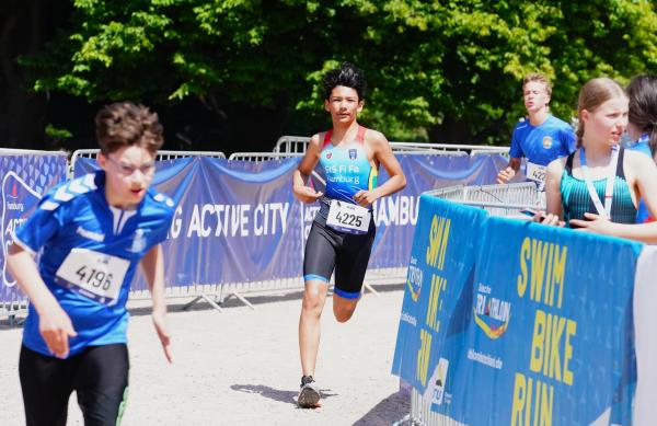 Zwei Jungs laufen um eine Kurve, an den Seiten sind Banner an den Absperrungen angebracht und Kinder stehen dahinter zum Anfeuern