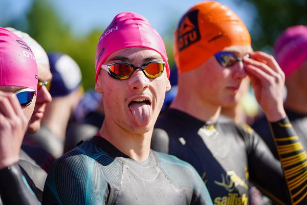 Jugendliche im Neo mit Badekappe und Schwimmbrille auf, einer von ihnen streckt du Zunge raus