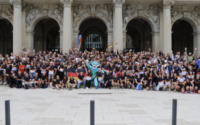 Viele Menschen in deutschen Trikots jubeln mit Fahnen für ein Gruppenbild - in der Mitte das Maskottchen der Deutschen Triathlon Union
