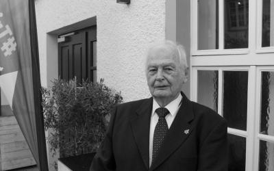 Ein älterer Mann steht vor einer Fensterfront und schaut in die Kamera