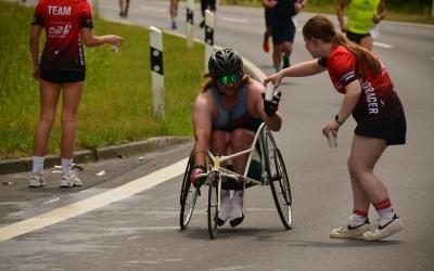 Ein Para Triathlet im Rollstuhl bekommt während seines Wettkampfs von einer Frau einen Trinkbecher gereicht