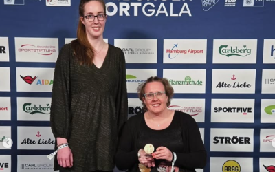 Eine Frau im Rollstuhl und eine Frau neben ihr stehend werden vor einer Sponsorenwand im Hintergrund portraitiert