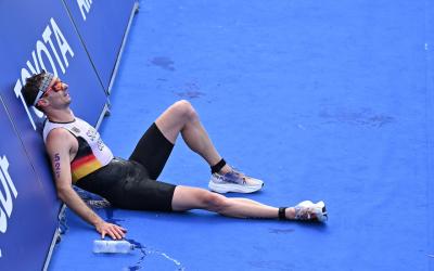 Ein deutscher Para Triathlet sitzt auf dem Boden im Zielbereich und lehnt sich erschöpft an eine Bande an