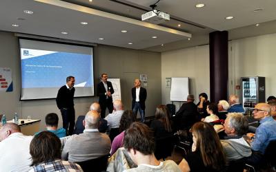 Drei Männer sprechen im Rahmen einer Präsentation zu vielen Menschen, die auf Stuhlreihen sitzen und zuhören