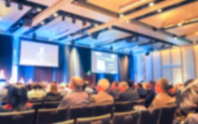 Mehrere Menschen sitzen bei einer Rede mit dem Rücken zur Kamera in einem Saal 