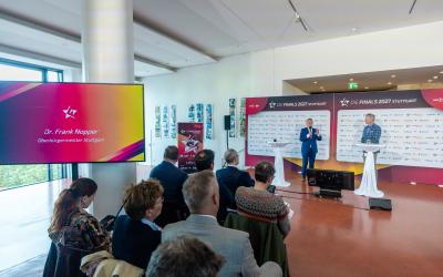 Zwei Männer unterhalten sich vor einer Presserückwand im Rahmen einer Pressekonferenz. Im Vordergrund sieht man mehrere Menschen, die auf Stühlen sitzend interessiert zuhören