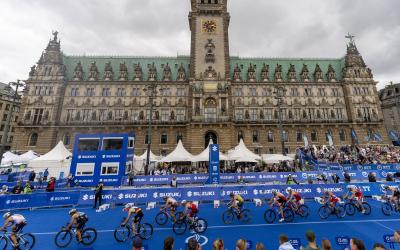 Zahlreiche Triathleten fahren auf dem Rad wie an einer Perlenschnur aufgereiht hintereinander durch die Wechselzone. Im Hintergrund ist das Hamburger Rathaus prominent zu sehen