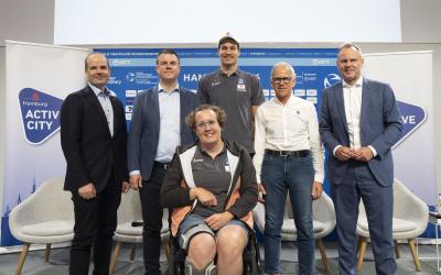 Fünf Männer posieren gemeinsam mit einer Rollstuhlfahererin für ein Foto auf der Bühne der Pressekonferenz